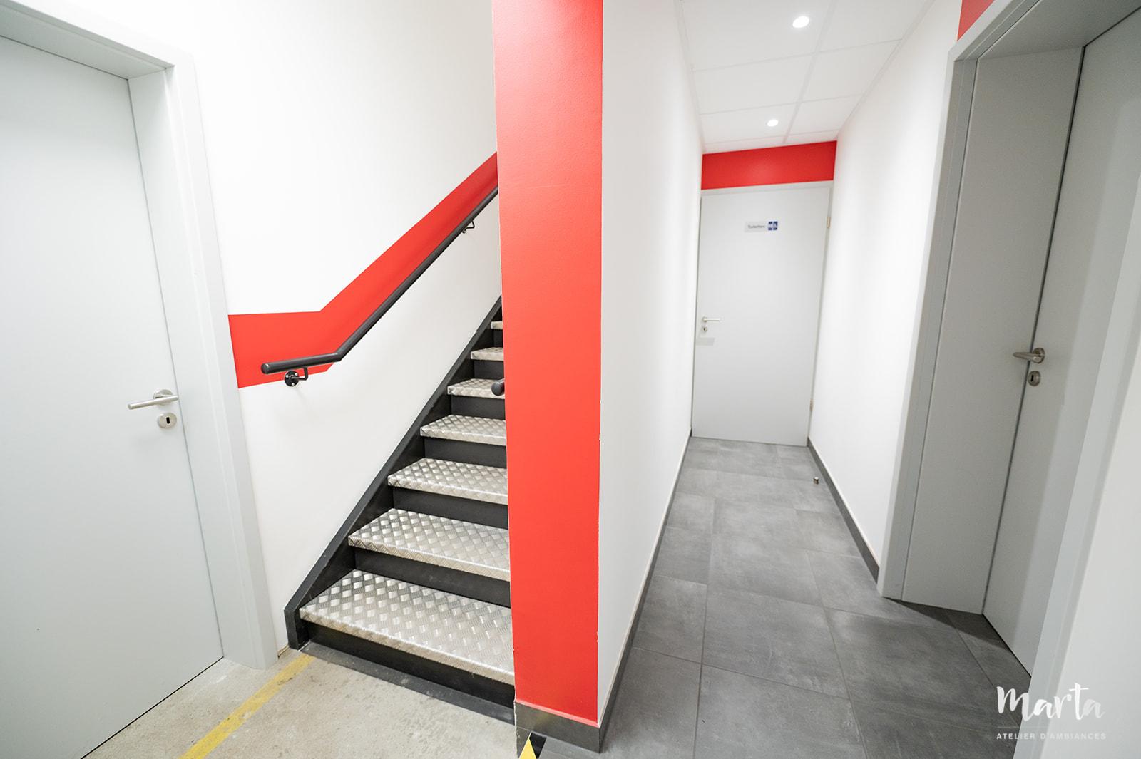Couloir modernisé avec carreaux gris foncé au sol, murs blancs et accents rouges, rappelant la couleur du logo de l'entreprise, créant une ambiance contemporaine et dynamique