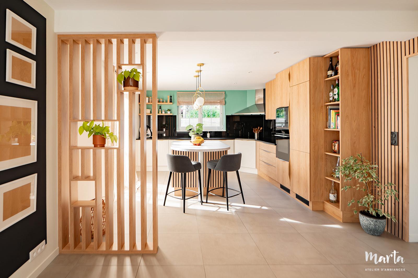 Cuisine contemporaine avec îlot central, meubles blancs et bois, plan de travail en granit noir, séparée par un claustra en bois. – Marta Atelier d’Ambiances